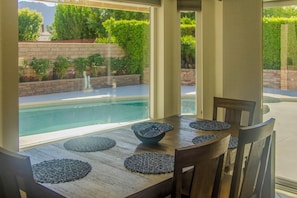 A dining table with placemats and chairs, overlooking a swimming pool with a view of a landscaped garden.