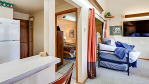 Un espacio habitable compacto con kitchenette, una cama con colchón azul y gris, y un televisor montado en la pared.