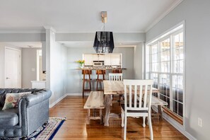 Un coin repas avec une table et des chaises en bois, une cuisine à l’arrière-plan et une grande fenêtre avec des stores.
