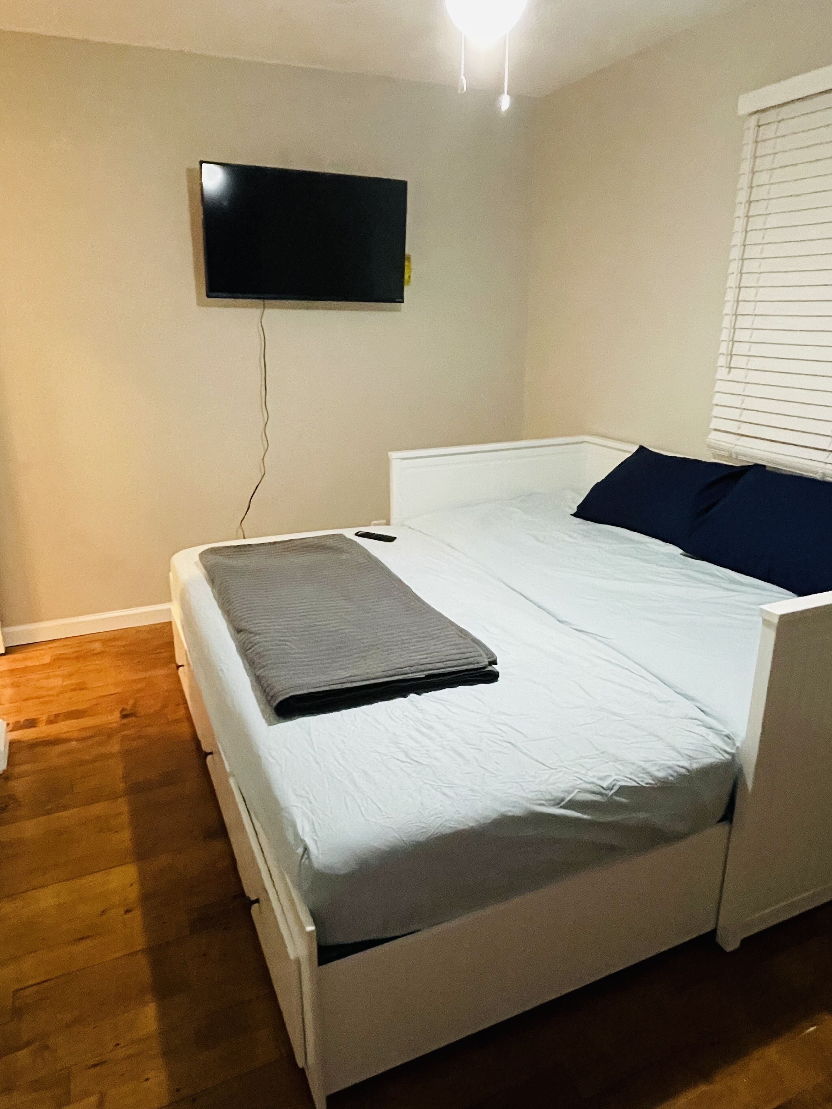 Habitación individual con una cama, televisión de pantalla plana en la pared, mesita de noche y ventana con persianas.