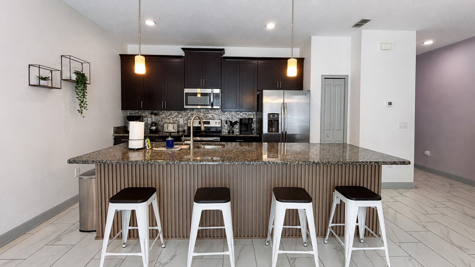 A modern kitchen with dark cabinets, stainless steel appliances, and a granite countertop. There are three white bar stools and pendant lights above the island.