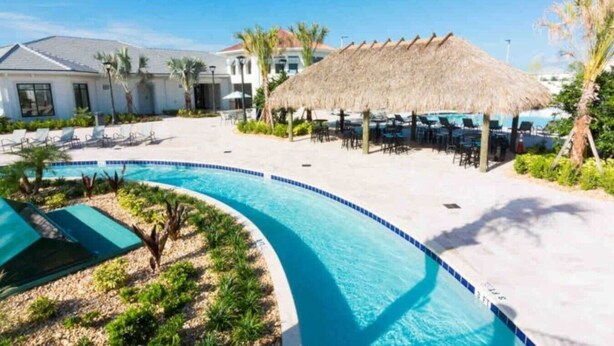 A pool area with a curved pool, lounge chairs, a thatched umbrella, and a building in the background.