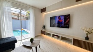 A modern living room with a flat-screen TV mounted on the wall, a pool visible through the glass door, and a small table with a plant.