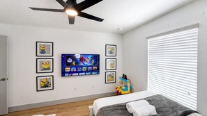 Un dormitorio con cama, ventilador de techo, televisor montado en la pared y ventana con persianas.