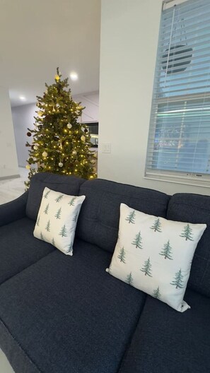 A dark-colored sofa with two pillows featuring a tree pattern, a Christmas tree with lights, and a window with blinds.