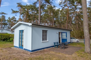 Un petit bâtiment d’un seul étage, avec une porte et des fenêtres bleues, une terrasse avec une table et des chaises, et une voiture garée à proximité.