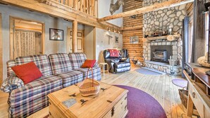 A cozy log cabin interior with a plaid sofa, a fireplace, and a stone wall. - Pet-Friendly New York Cabin w/ Hot Tub & Game Room (Maplecrest)