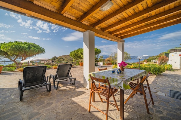 An outdoor patio with stone flooring, wooden chairs, a table with a tablecloth, and a view of the sea and hills.
