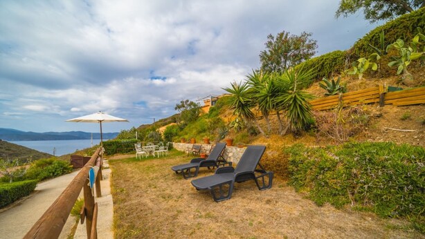 Um pátio com cadeiras de descanso, uma mesa e um guarda-sol, com vista para uma encosta arborizada e um corpo d'água ao longe.