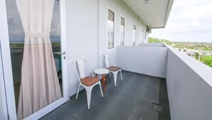 Un balcon avec deux chaises blanches et une petite table, une porte vitrée avec des rideaux, et une vue sur un paysage verdoyant.
