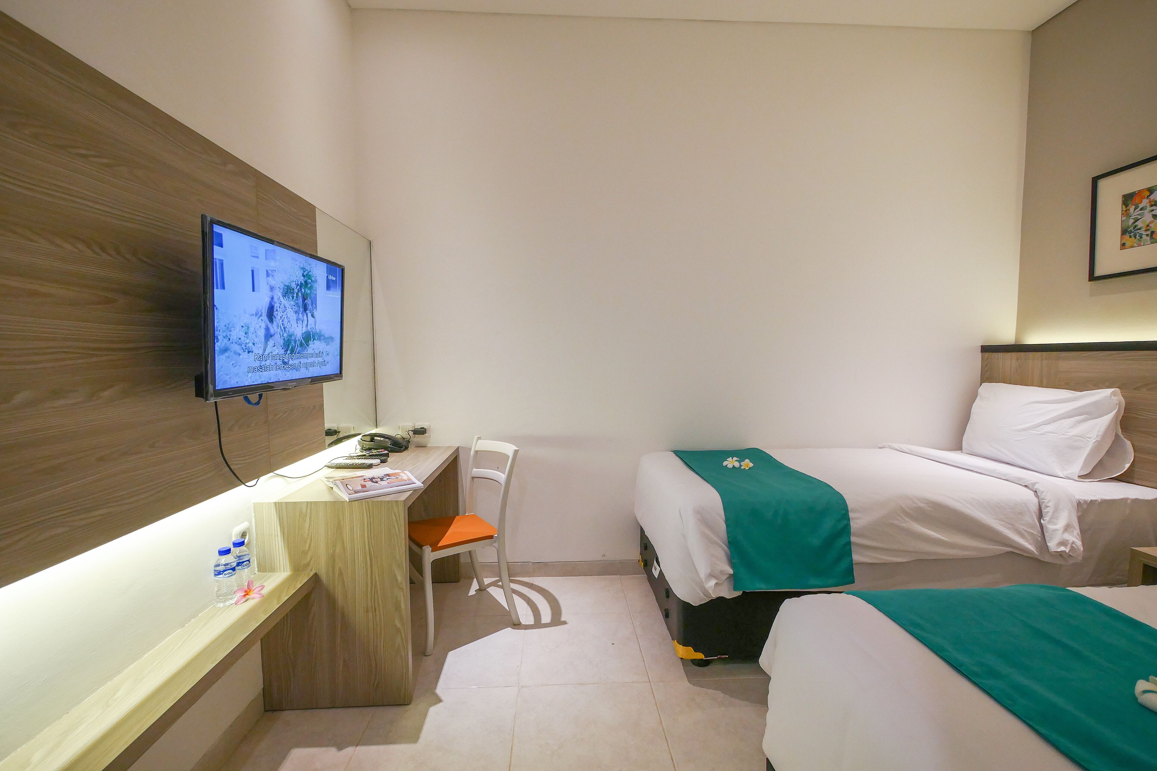 A hotel room with a wooden desk, a chair, a bed with a green blanket, and a TV mounted on the wall.
