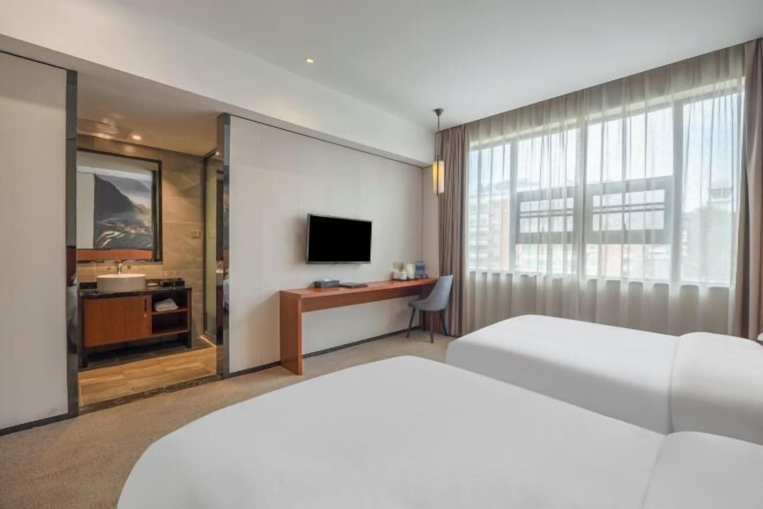 Une chambre d’hôtel avec un grand lit, un bureau avec une chaise, une télévision, une salle de bain avec un miroir et une fenêtre avec des rideaux.