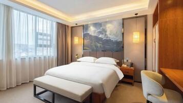 A hotel room with a large bed, a desk, a chair, a painting on the wall, and a window with curtains.