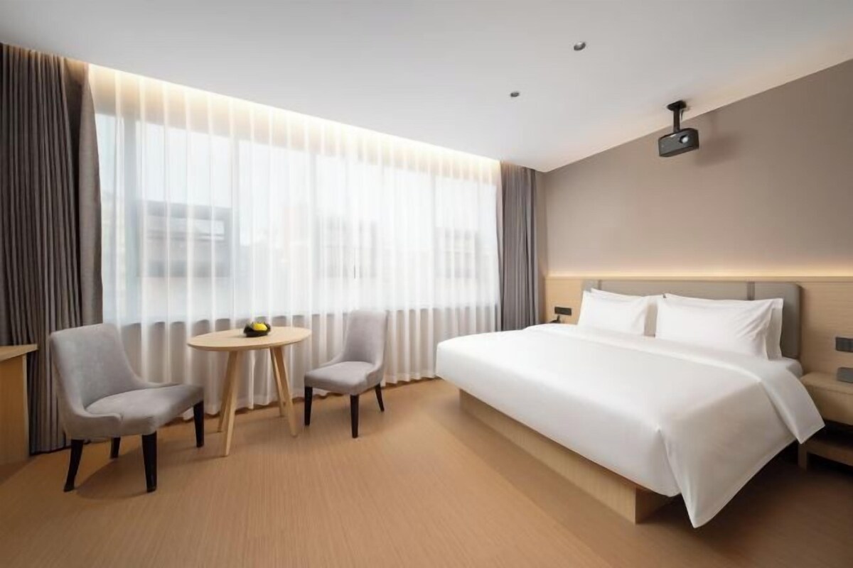 A hotel room with a large bed, two chairs, a small table, and a window with curtains.
