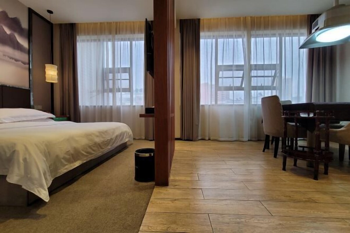 A hotel room with a bed, a desk, a chair, a lamp, and a view of the city through large windows.