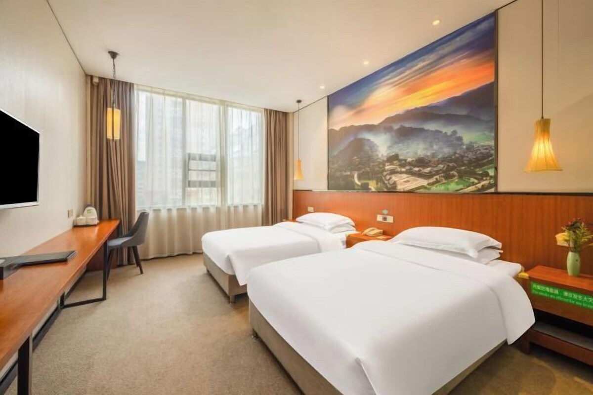 A hotel room with two beds, a desk, a TV, and a painting on the wall.