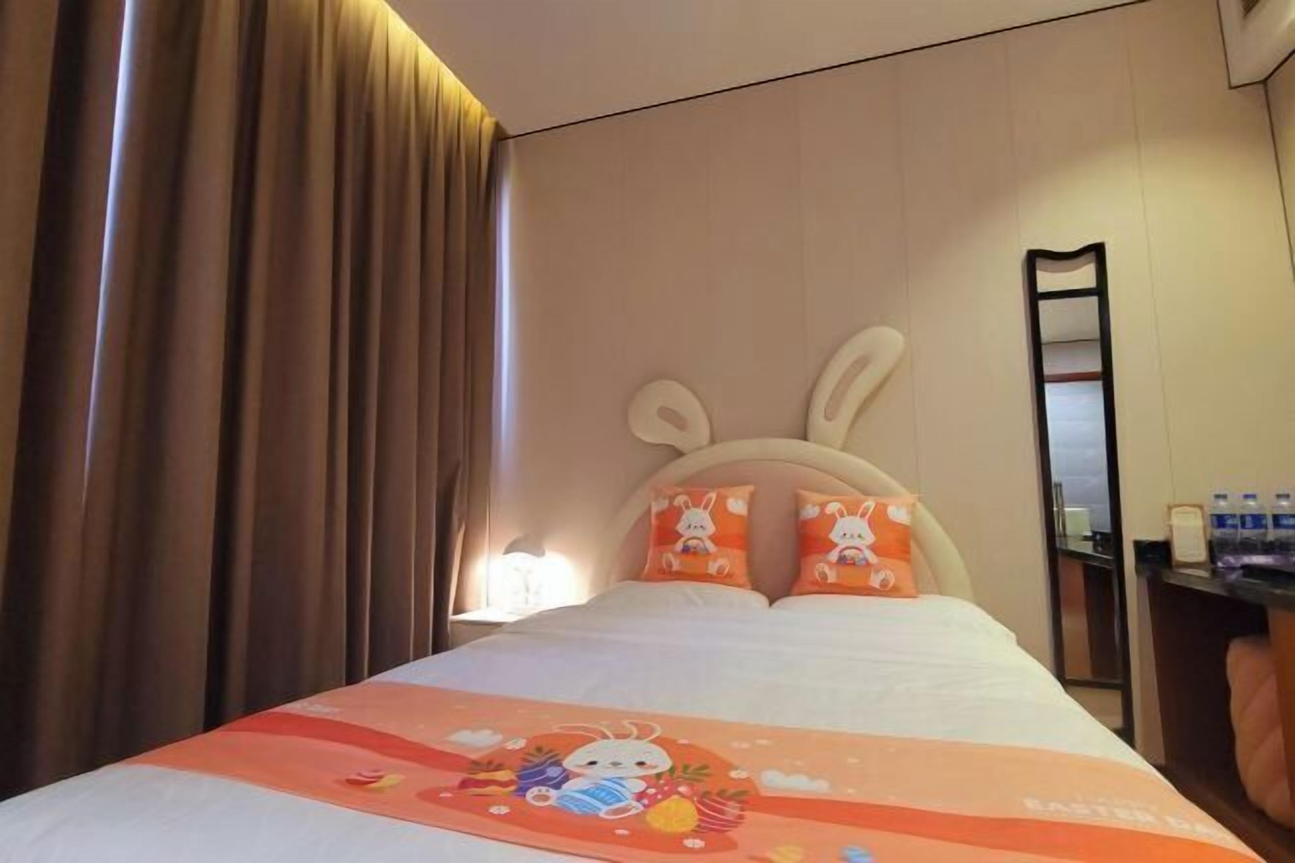 A hotel room with a bed, curtains, a headboard with bunny pillows, a bedside table with a lamp, and a mirror.