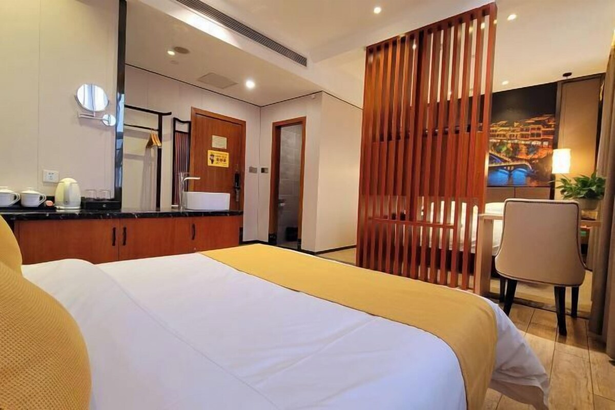 A hotel room with a bed, a desk, a chair, a mirror, and a view of a cityscape.