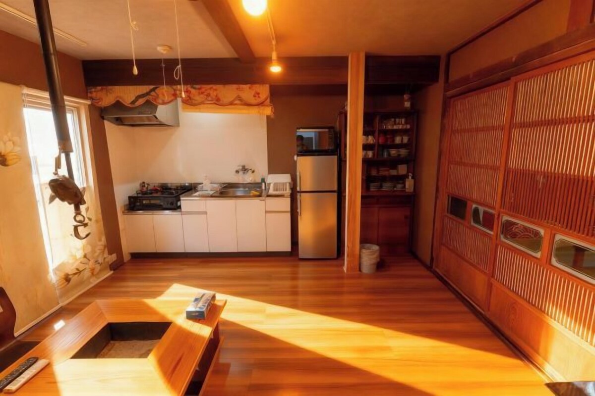 Cocina compacta con refrigerador, microondas y gabinetes blancos. Incluye un banco de madera y una pequeña mesa con un libro.