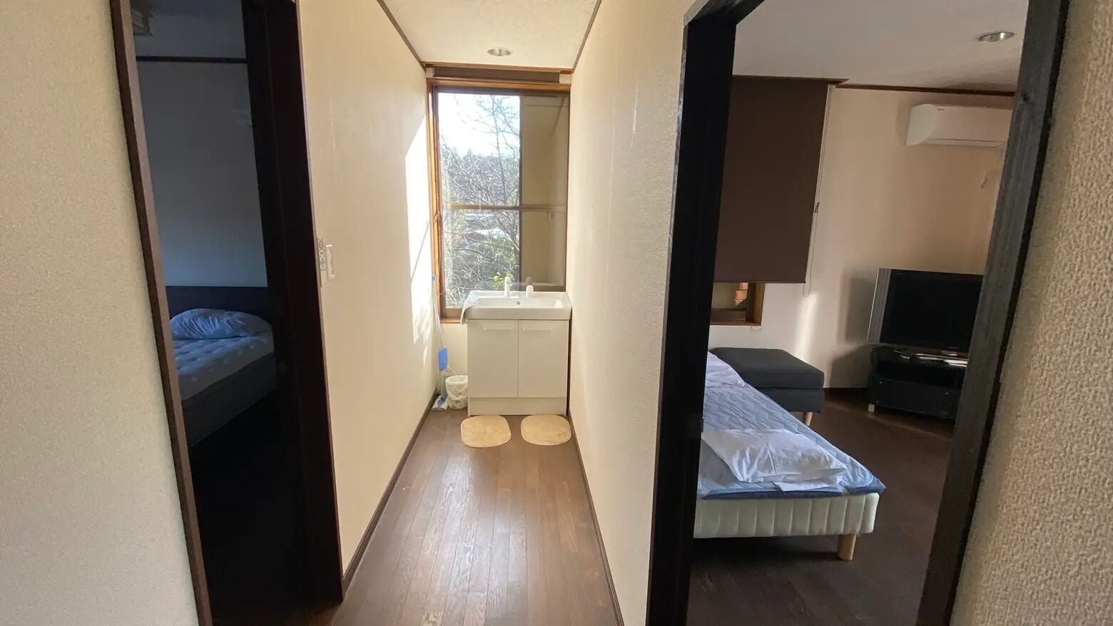 A narrow hallway leading to a bedroom with a bed, a small window, and a bathroom with a sink.