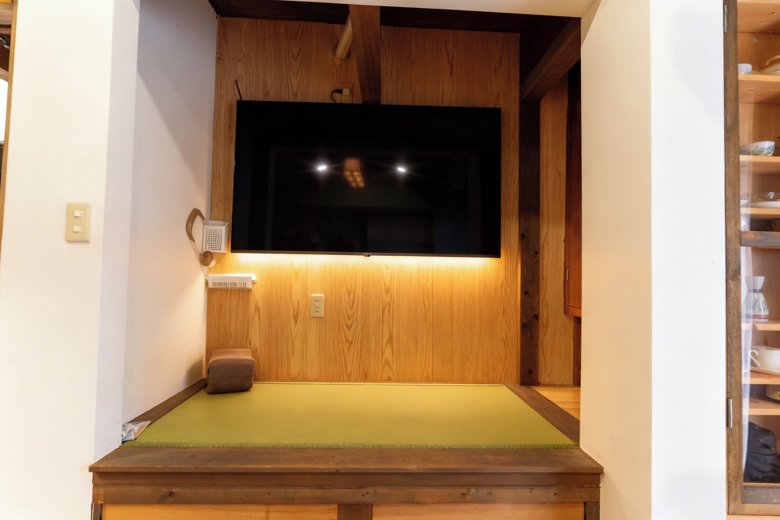 Une pièce avec un sol recouvert de tatamis, un plafond en bois, une télévision et une étagère où sont posées des tasses et des bols.
