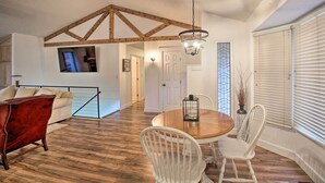 Een eethoek met een houten tafel en witte stoelen, een rode stoel, een hanglamp en uitzicht op de keuken door een open doorgang.