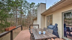 Een houten terras met een tafel, stoelen, een barbecue en een raam met uitzicht op een bebost gebied.