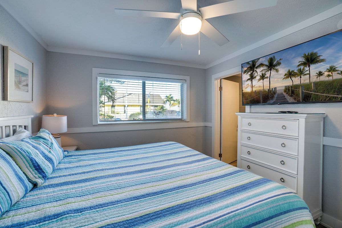 Een slaapkamer met een bed, een plafondventilator, een raam met jaloezieën, een dressoir en een schilderij van een strandgezicht.