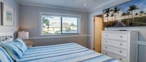 A bedroom with a bed, a ceiling fan, a window with blinds, a dresser, and a painting of a beach scene.