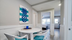 Un comedor con una mesa blanca y sillas de mimbre, una obra de arte con forma de piña azul en la pared, y vista a la habitación contigua.