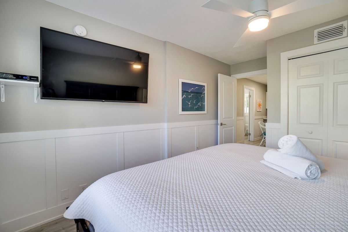 A hotel room with a bed, a flat-screen TV mounted on the wall, a ceiling fan, and a door leading to a bathroom.