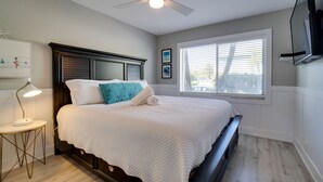 Un dormitorio con cama, ventilador de techo, mesita de noche, lámpara, ventana con persianas y televisor instalado en la pared.