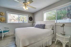 Un dormitorio con cama, ventilador de techo, ventana con persianas, una silla y una pequeña mesa con lámpara.