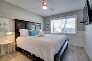 A bedroom with a bed, a ceiling fan, a window with blinds, and a wall-mounted TV.