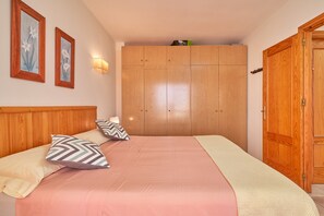 Een slaapkamer met een houten hoofdeinde, een bed met roze en witte beddengoed, twee wandlampen en een houten garderobe.