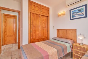 A bedroom with a wooden wardrobe, a bed with a striped bedspread, a bedside lamp, and a wall-mounted air conditioner.