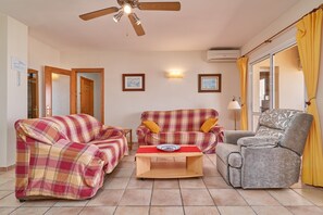 Een woonkamer met een plafondventilator, twee banken, een salontafel en een grijze fauteuil.