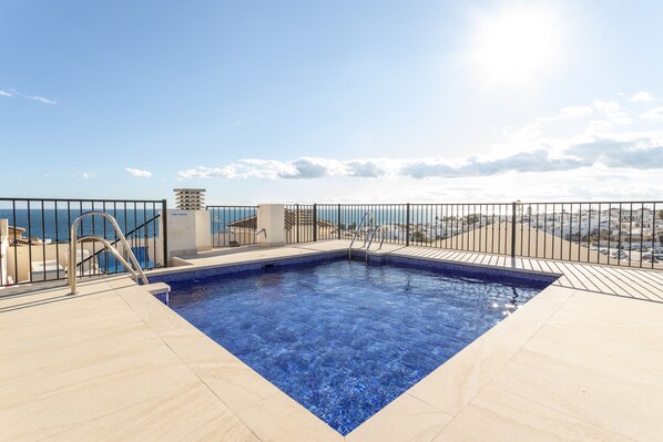 A rooftop pool with a view of the sea, surrounded by a metal fence and a tiled deck.