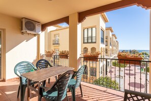 A balcony with a table and chairs, overlooking a courtyard with buildings and a view of the sea.