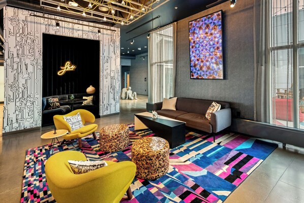 A modern lounge area with a sofa, armchairs, and ottomans, featuring geometric patterned rugs and a large abstract painting. - Aloft by Marriott Anchorage (Anchorage)
