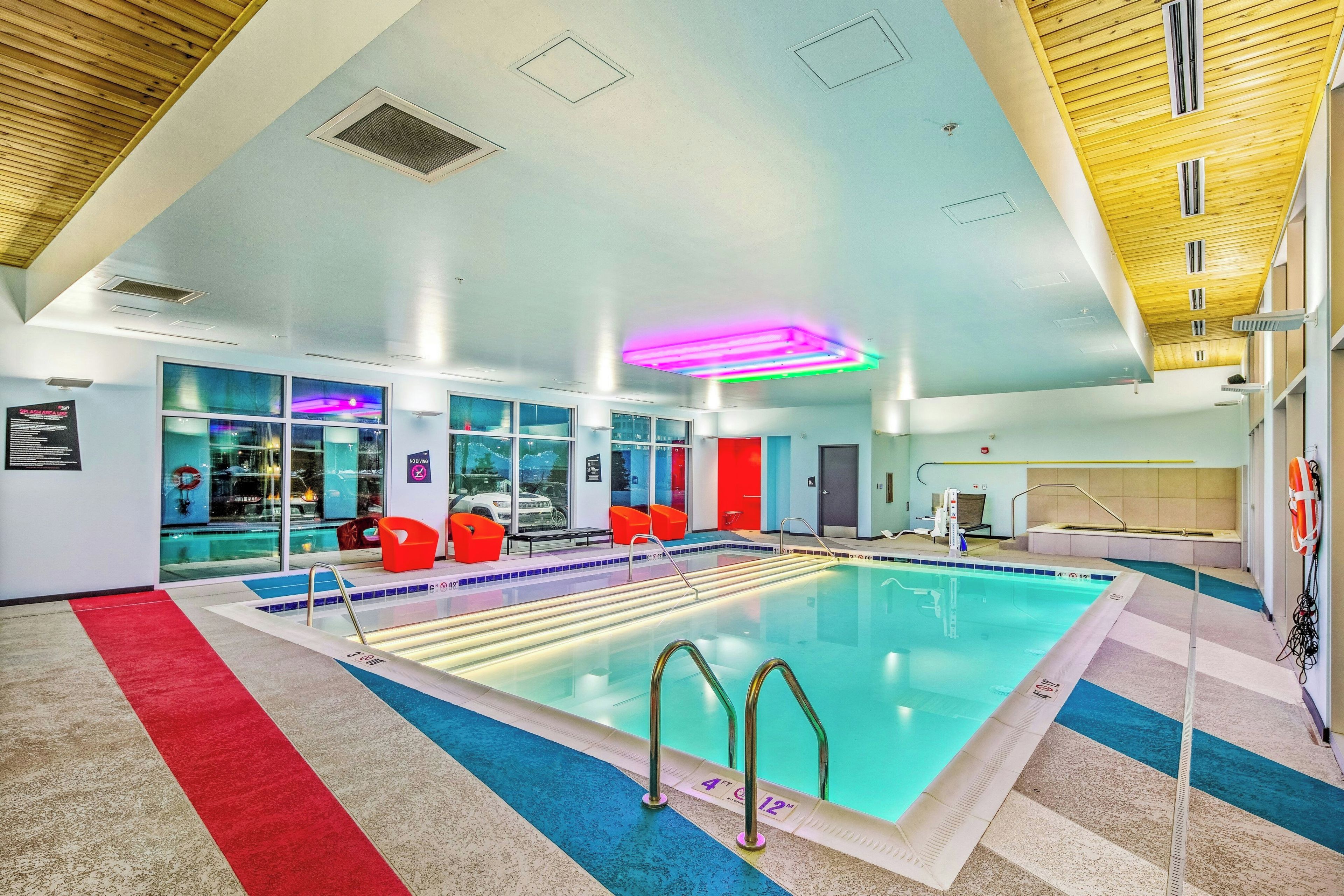 An indoor swimming pool with clear water, a red and blue tiled floor, and a ceiling with wooden panels and recessed lighting.