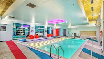 An indoor swimming pool with clear water, a red and blue tiled floor, and a ceiling with wooden panels and recessed lighting.
