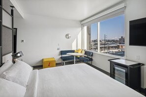 A hotel room with a bed, a small table, a chair, a TV, and a window with a city view.