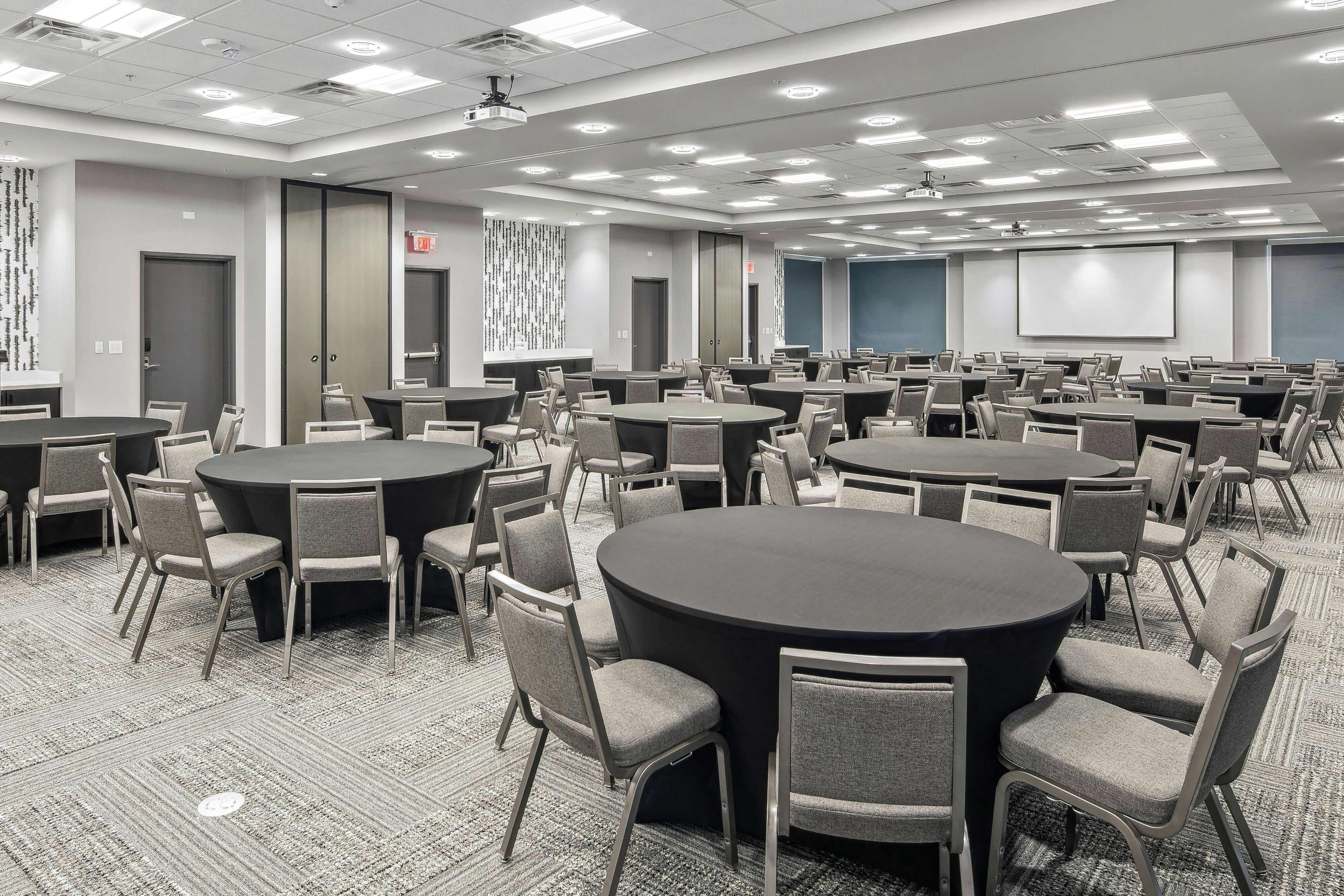 Sala de conferências com várias mesas redondas e cadeiras, equipada com projetor e tela.