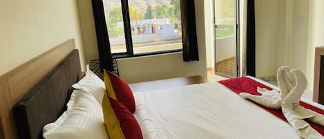 A neatly made bed with white and red pillows, a wooden headboard, and a view of a mountainous landscape through a window with black curtains.