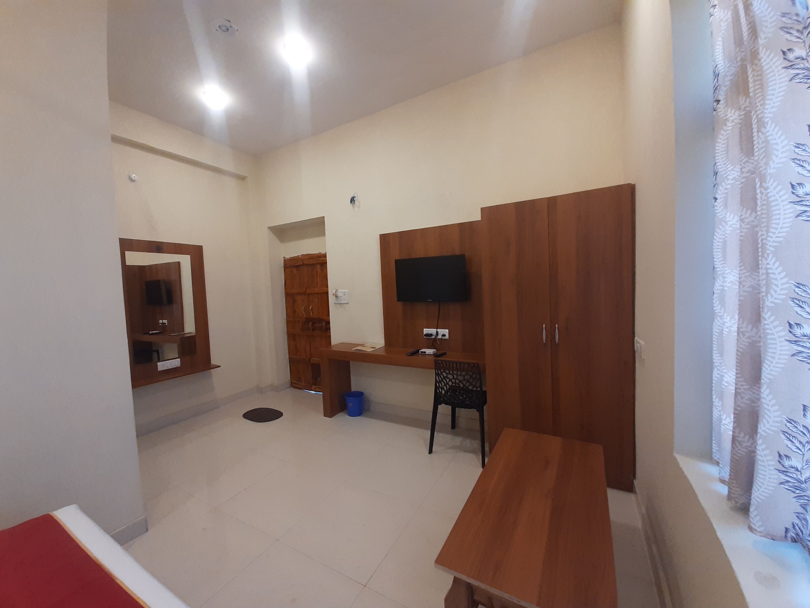 Una habitación con un armario de madera, un escritorio, una silla, un televisor montado en la pared y una ventana con cortina.