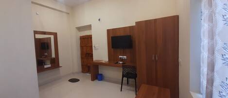 Una habitación con un armario de madera, un escritorio, una silla, un televisor montado en la pared y una ventana con cortina.