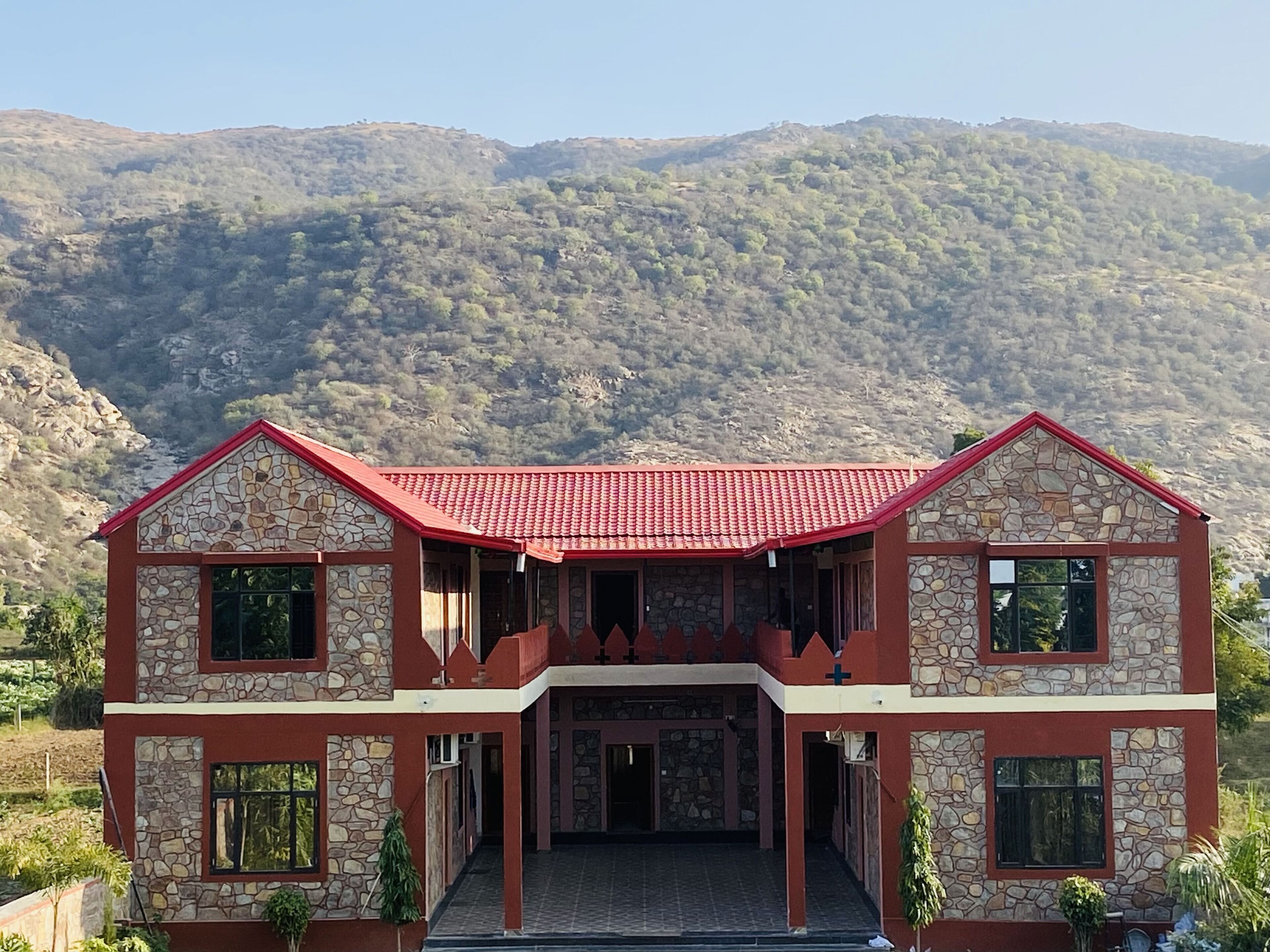 A two-story building with a red roof and stone facade, set against a mountainous backdrop.