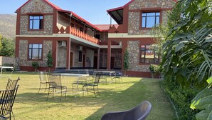 A two-story building with stone and red exterior walls, a covered outdoor seating area with metal chairs, and a well-maintained lawn. - Pushkar Retreat Resort (Pushkar)