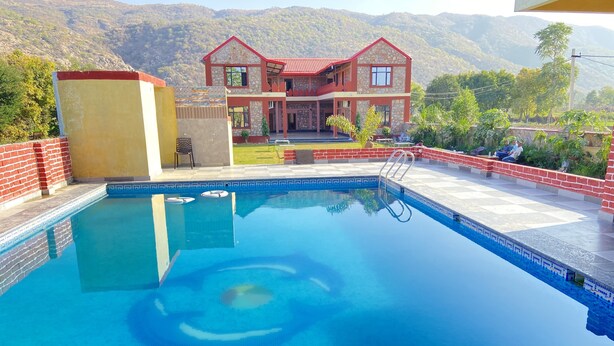 A swimming pool with a clear view of a two-story building with a red roof and stone facade in the background. - Pushkar Retreat Resort (Pushkar)